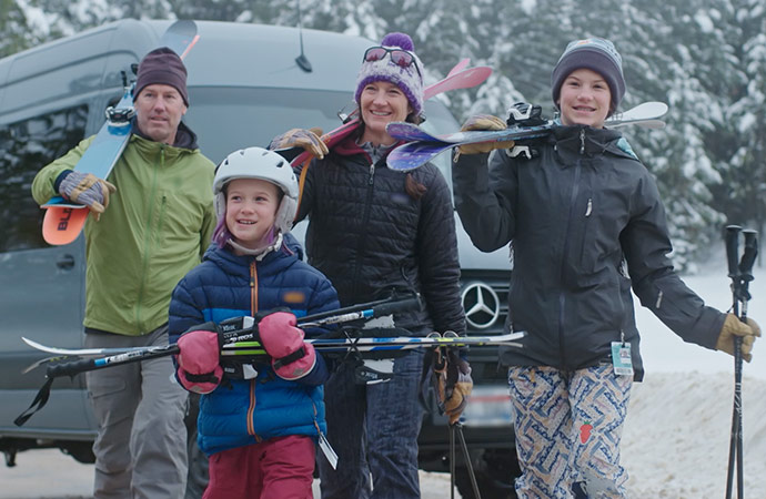 Clickable video still of family with ski equipment in the mountains