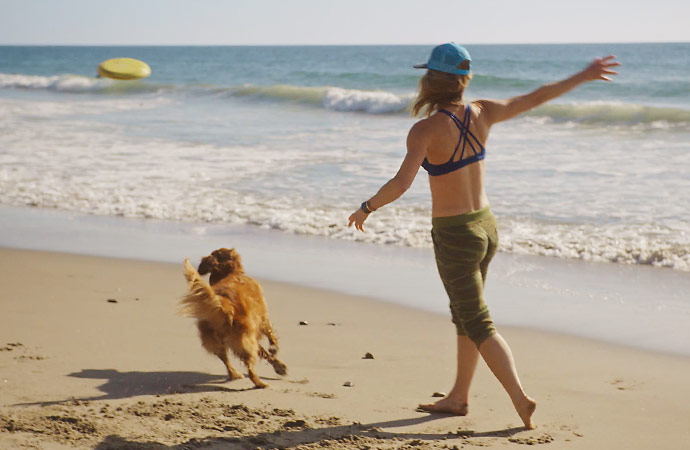 Clickable video still of woman throwing frisbee with dog at the beach.